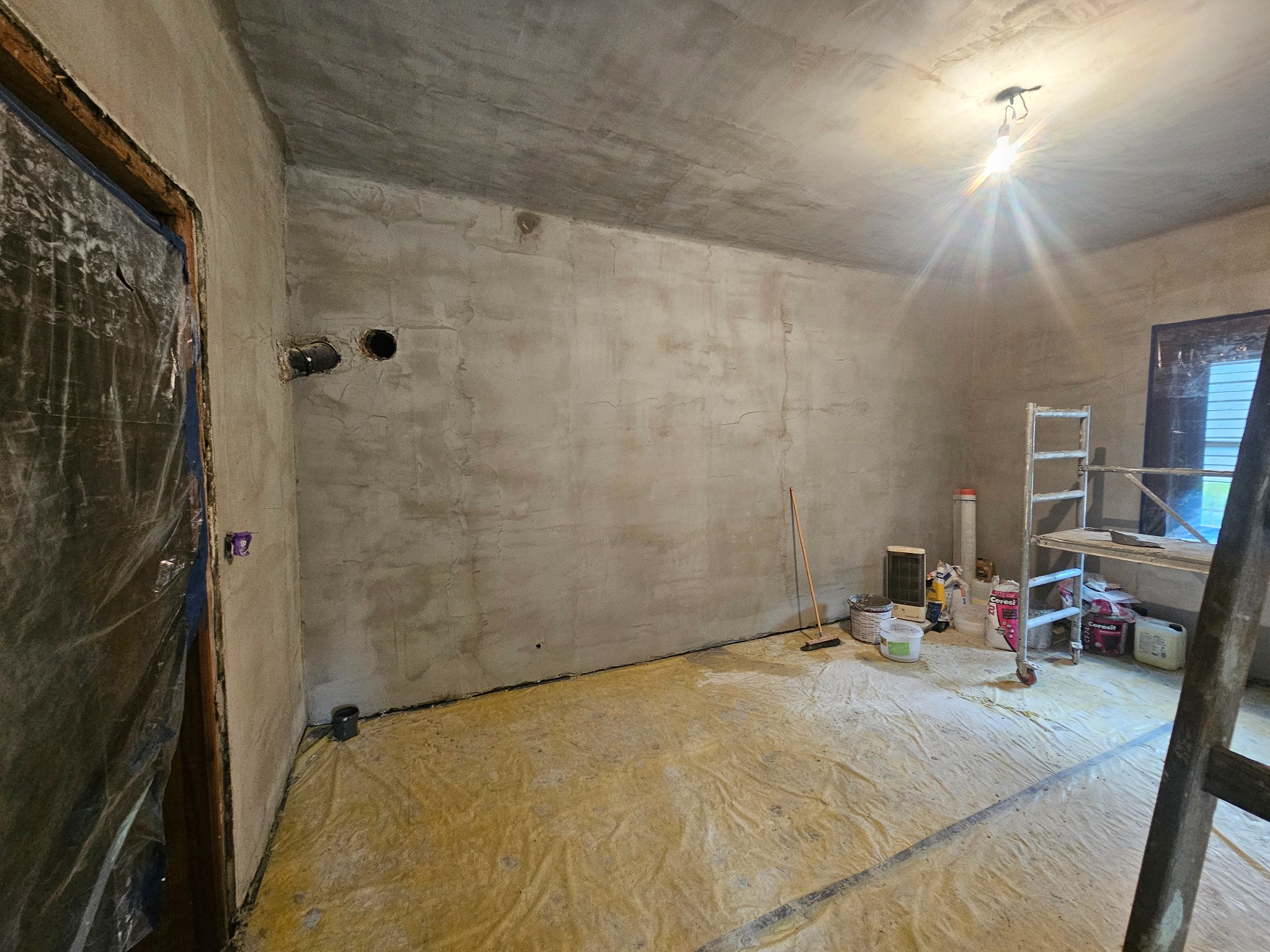 Pokój w trakcie prac wykończeniowych: surowe ściany po szpachlowaniu, zabezpieczona folią podłoga, drabina i narzędzia malarskie. Widoczne otwory w ścianie.