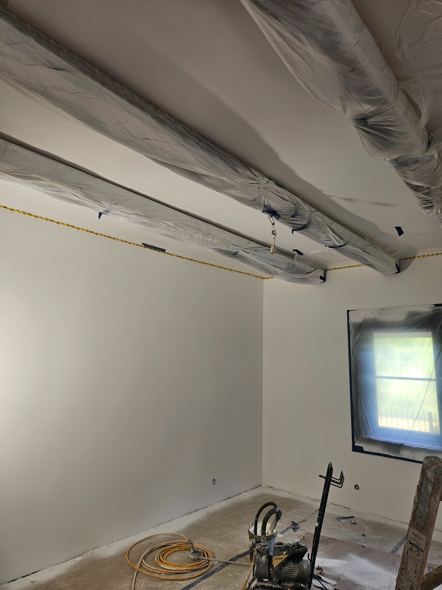 Białe ściany po malowaniu natryskowym, zabezpieczone folią sufit i okno, widoczny agregat malarski i drabina. Świeżo pomalowane wnętrze.