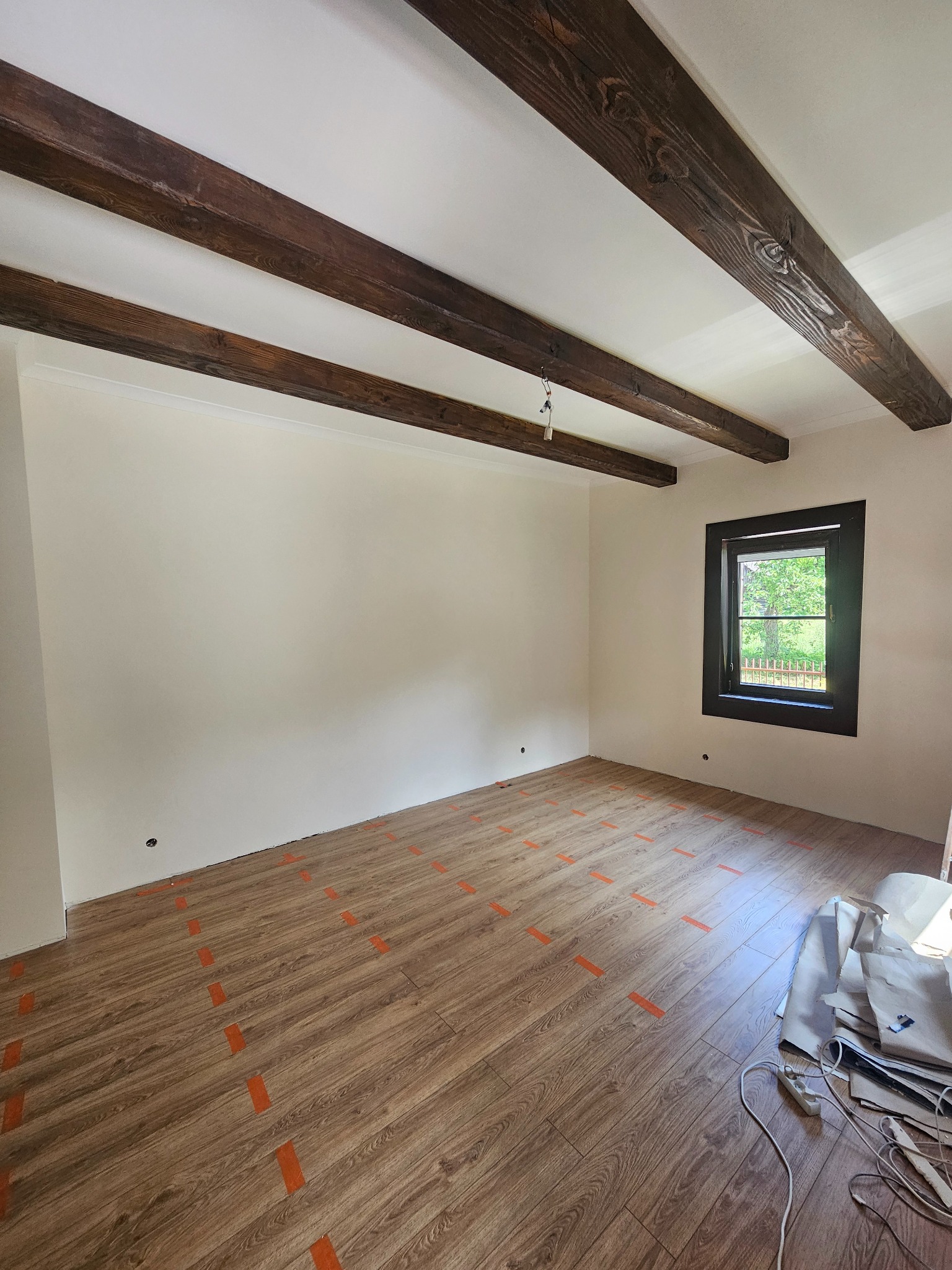 Jasne wnętrze z drewnianymi belkami stropowymi i panelami podłogowymi zabezpieczonymi taśmą. Okno z widokiem na zieleń. Przygotowanie do prac wykończeniowych.