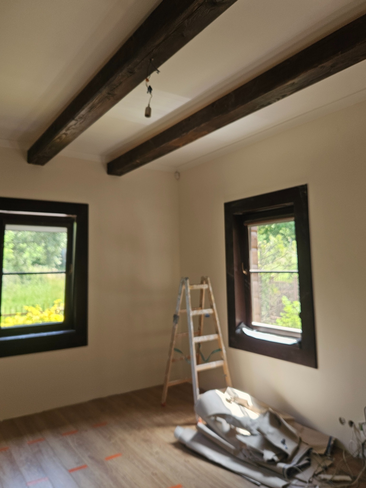 Pokój po malowaniu ścian na beżowo z drewnianymi belkami stropowymi i oknami w ciemnych ramach. Widoczna drabina i zabezpieczenia podłogi.