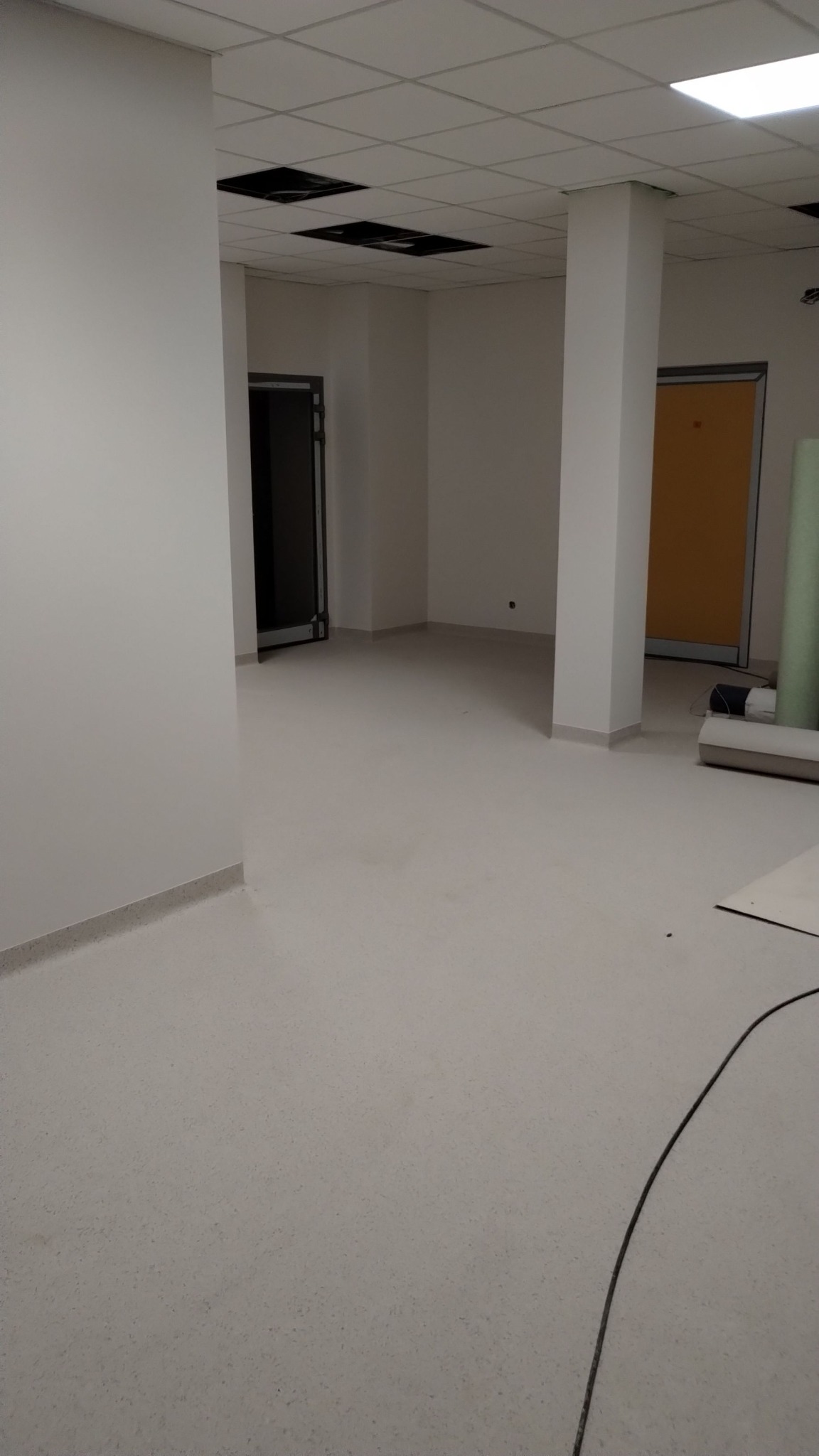 Wnętrze biurowe po remoncie: jasne ściany, podłoga z terakoty, sufit podwieszany z widocznymi otworami. Widoczne drzwi i kolumna. Minimalistyczny styl.