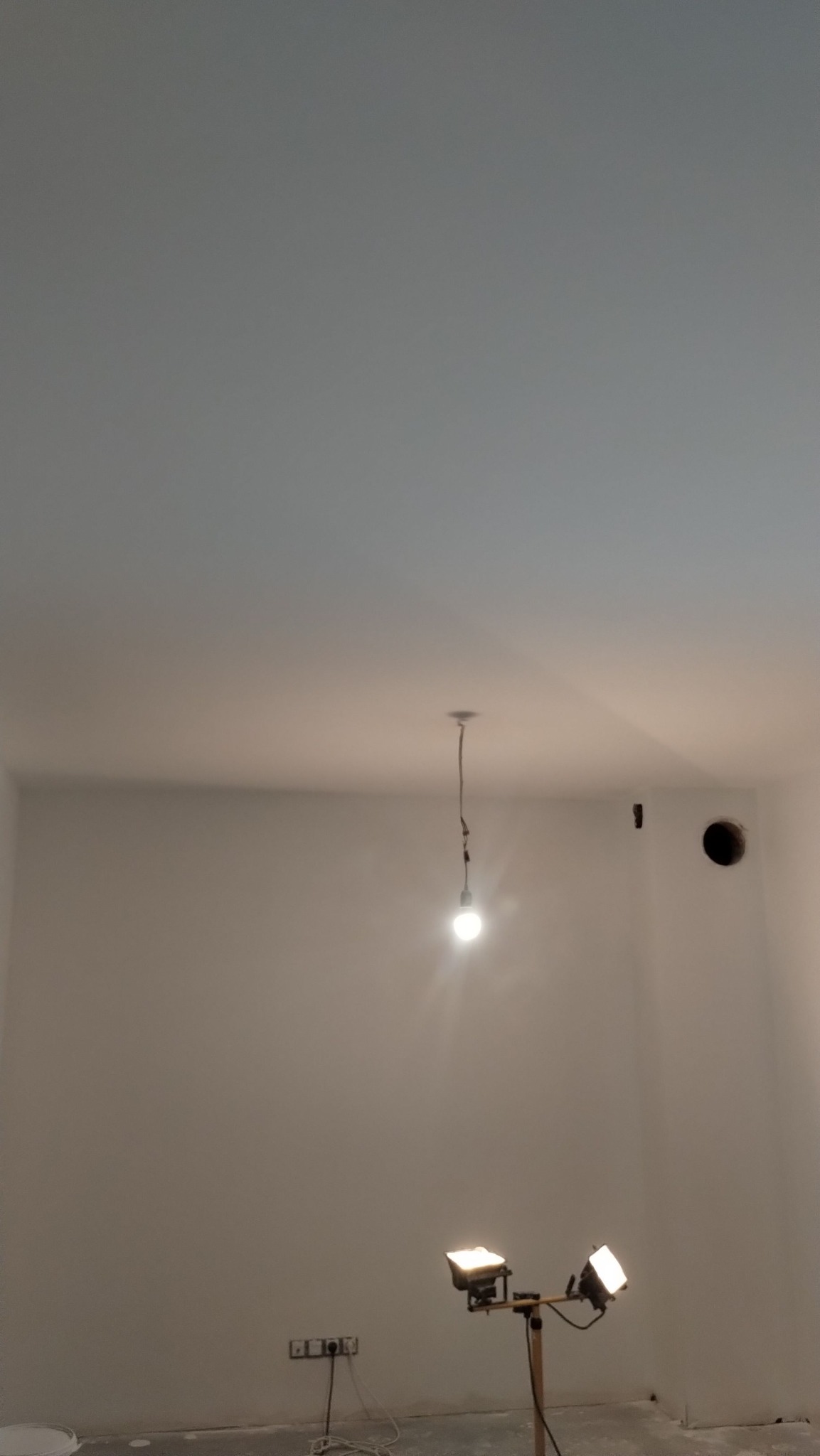 Wnętrze pokoju po szpachlowaniu ścian, widoczne gładkie, białe ściany, lampa wisząca i halogeny oświetlające. Przy podłodze widoczne gniazdka elektryczne.