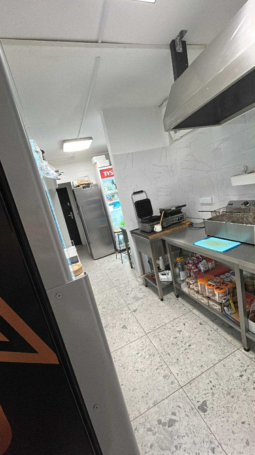 Wnętrze kuchni lokalu gastronomicznego z wyposażeniem: frytownica, gofrownica, stalowe stoły z zapasami. Widok na chłodziarkę i marmurowe płytki podłogowe.