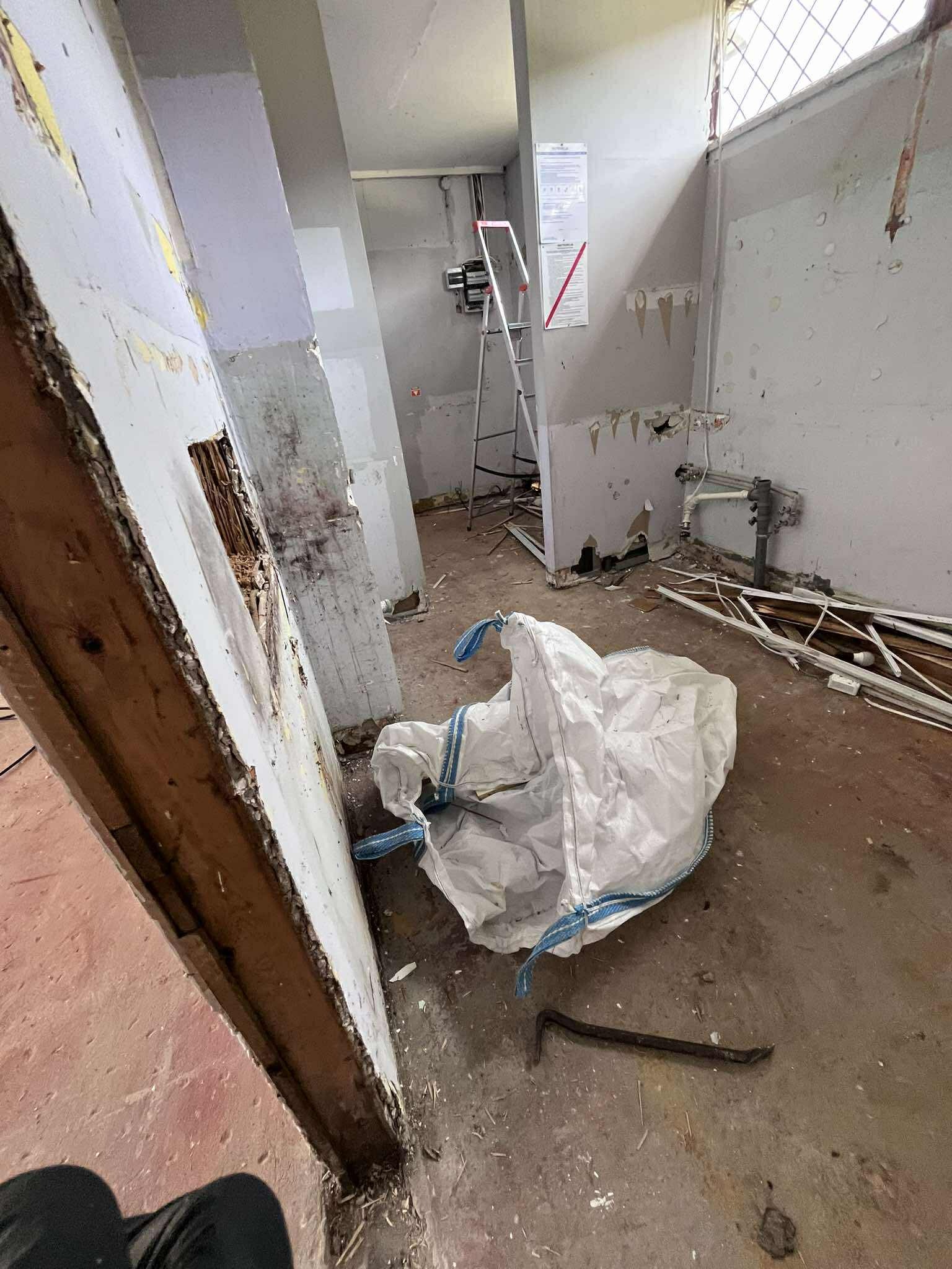 Wnętrze w trakcie demontażu: odsłonięte ściany, gruz w worku typu big bag, podważka na podłodze. Widoczne instalacje i drabina. Etap przygotowania do remontu.