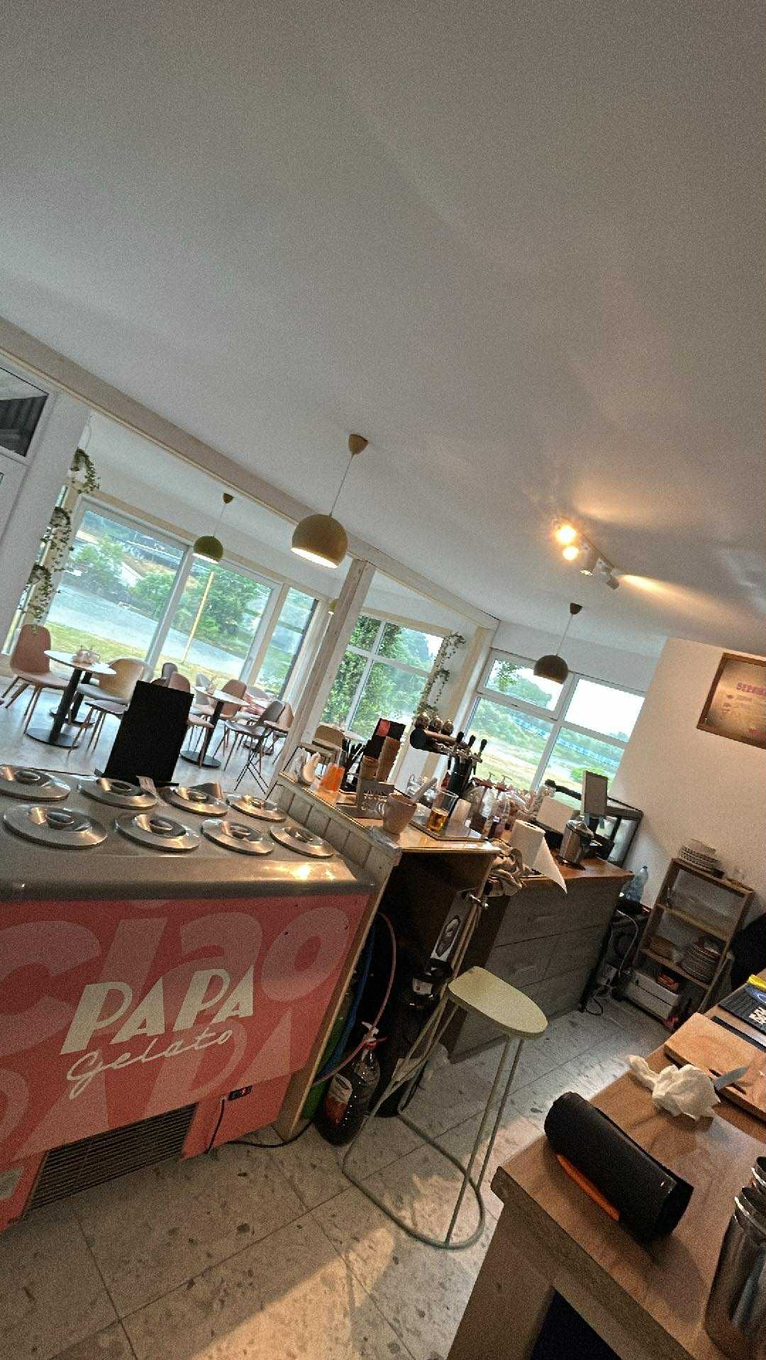 Jasne wnętrze kawiarni z widokiem na zewnątrz przez duże okna. Widoczny bar z lodami 'Papa Gelato', stoliki i krzesła dla gości. Oświetlenie punktowe i wiszące lampy.