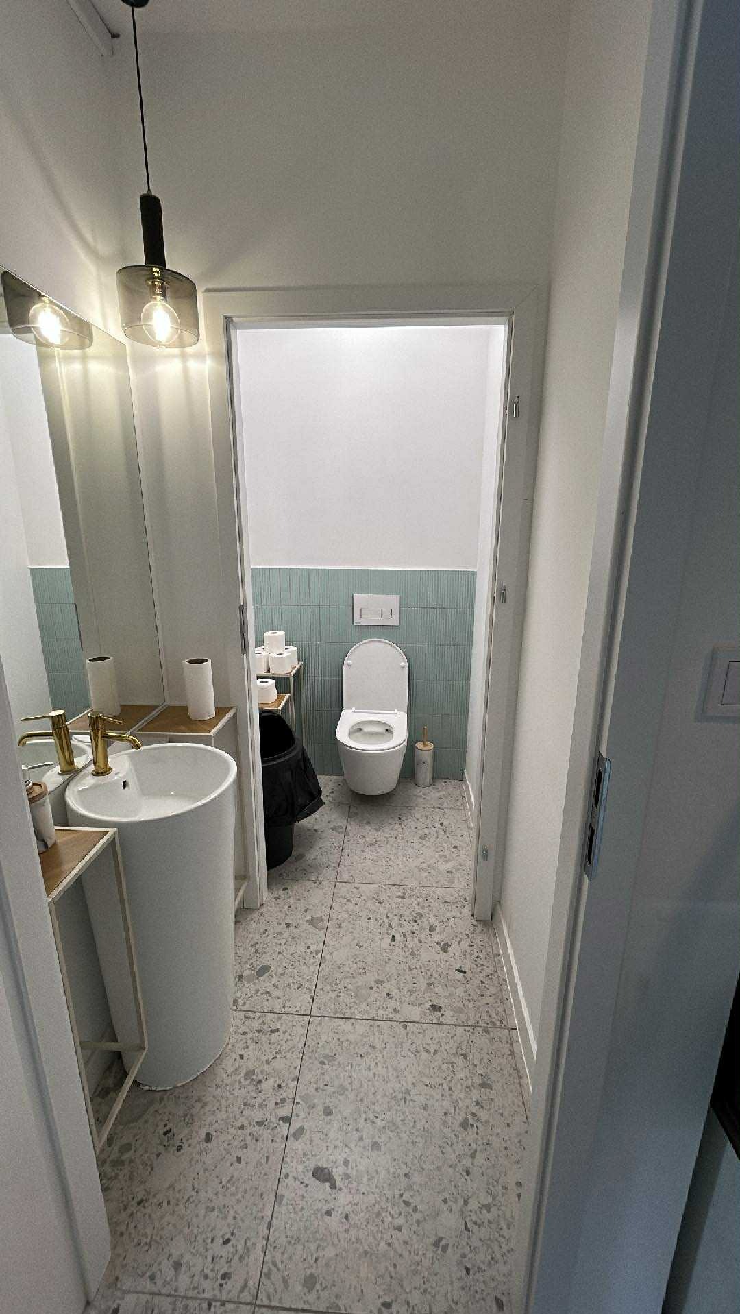 Nowoczesna łazienka z umywalką na postumencie, płytkami w odcieniach szarości i błękitu. Widok z korytarza, oświetlenie punktowe i minimalistyczny design.