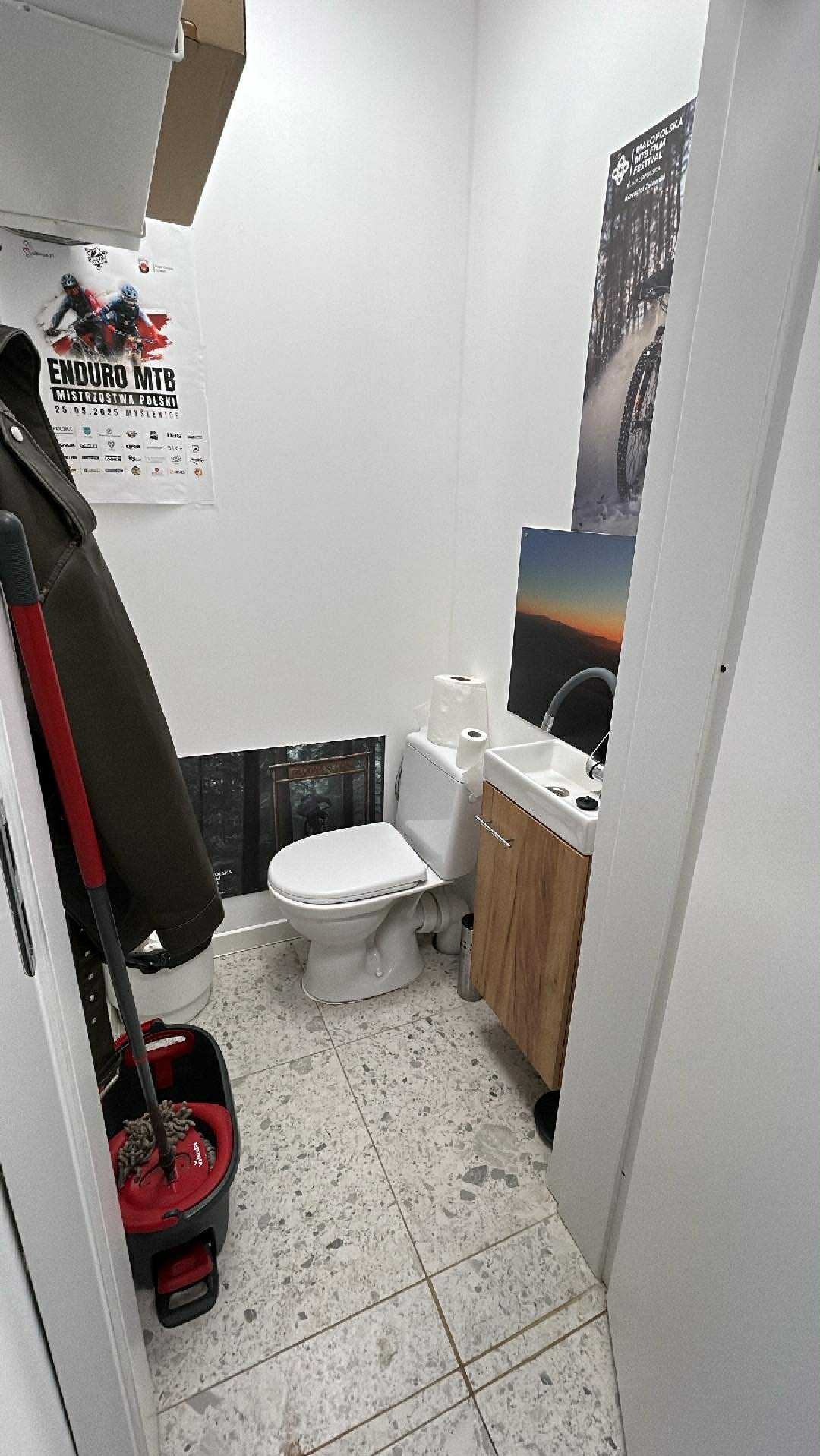 Mała łazienka z toaletą, umywalką z szafką i mopem. Na ścianach plakaty i grafiki. Podłoga wyłożona płytkami imitującymi lastryko.