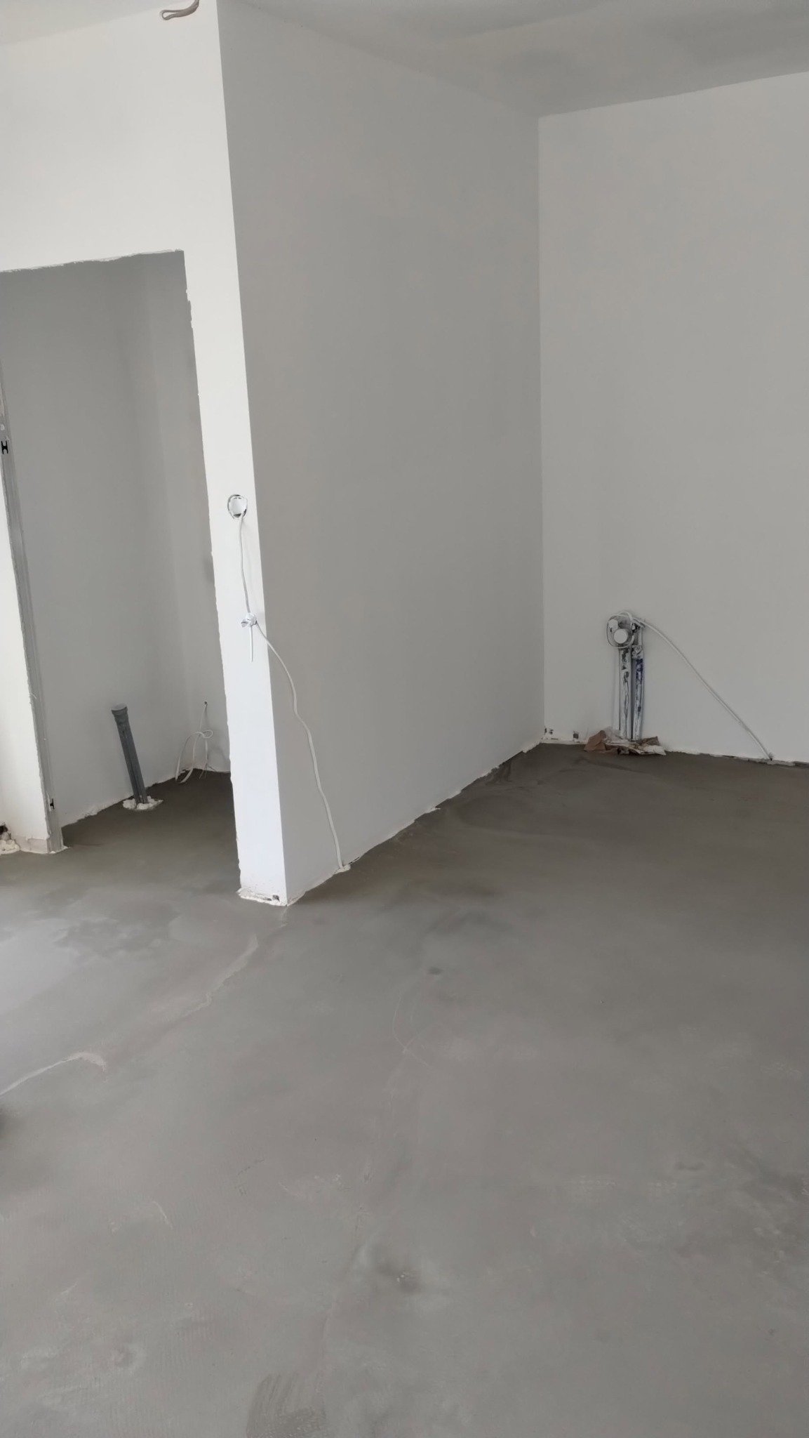 Wnętrze w trakcie prac wykończeniowych: świeżo wylana posadzka, białe ściany z wystającymi przewodami i rurami. Widoczne przygotowania do dalszych prac.