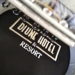 KIS PLUS KRZYSZTOF KAPUSTA ŁUKASZ KAPUSTA SPÓŁKA CYWILNA - Czarna czapka z białym haftem logo 'Divine Hotel & Resort' w trakcie procesu haftowania, widoczne metalowe elementy maszyny hafciarskiej.