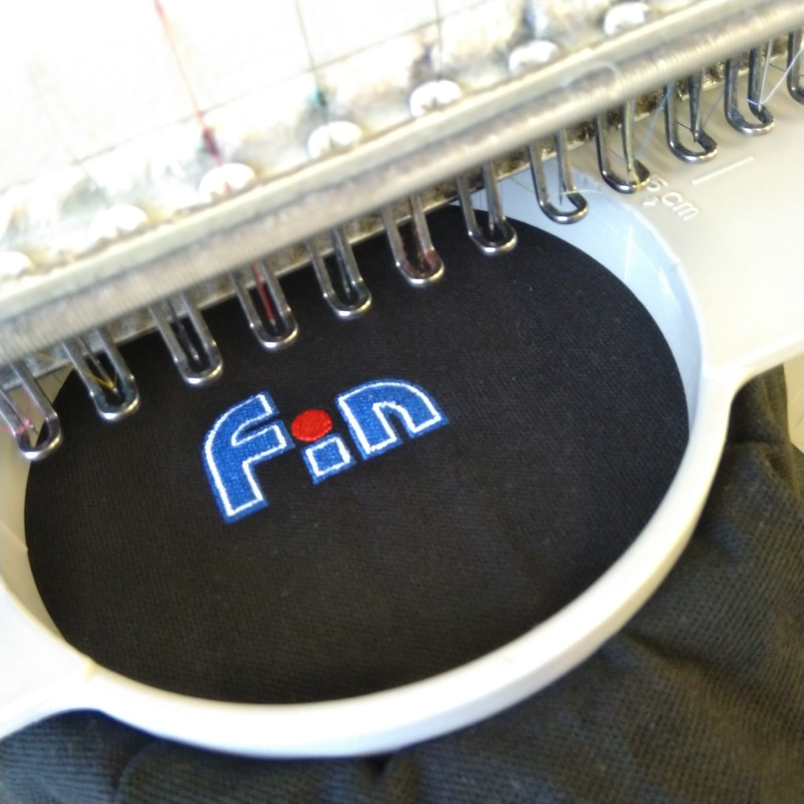 Zbliżenie na proces haftowania logo 'F.n' w kolorze niebieskim i czerwonym na czarnym materiale w maszynie do haftowania.