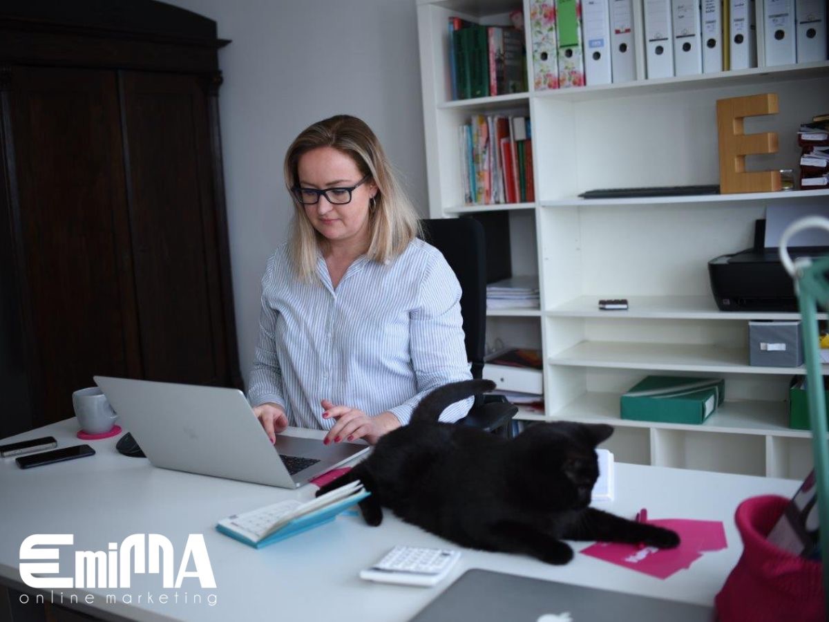 Kobieta w okularach pracuje na laptopie przy biurku z czarnym kotem, w tle regał z książkami i szafa.