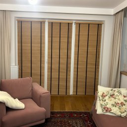 Growindo Dominik Tuszyński - Salon z drewnianymi żaluzjami w oknach. Widoczne beżowe zasłony, różowy fotel i kanapa. Na parapecie rośliny doniczkowe. Ciepłe oświetlenie wnętrza.