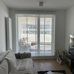 Growindo Dominik Tuszyński - Jasne wnętrze z białymi żaluzjami poziomymi, przez które widać balkon z meblami wypoczynkowymi. Minimalistyczny design, naturalne światło.