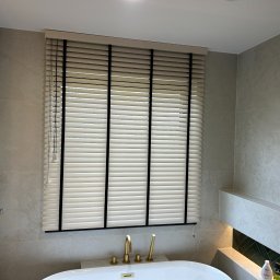 Growindo Dominik Tuszyński - Beżowe poziome lamele z czarnymi taśmami w łazience z wanną i złotą armaturą. Stonowana kolorystyka płytek i oświetlenie tworzą spójny, elegancki wystrój.