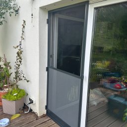 Growindo Dominik Tuszyński - Szara moskitiera ramkowa zamontowana przy drzwiach tarasowych, widok z zewnątrz na drewniany taras i roślinność w tle. Detal: widoczne zawiasy i uchwyt moskitiery.