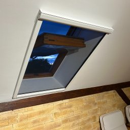 Growindo Dominik Tuszyński - Okno dachowe z moskitierą w białej ramie, widok na wieczorne niebo. W tle ściana z kamienia dekoracyjnego i fragment mebli. Ujęcie od dołu, podkreślające montaż.