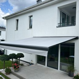Growindo Dominik Tuszyński - Biała markiza tarasowa z czarnym obramowaniem, zamontowana przy nowoczesnym domu z ogrodem. Widoczne rośliny ozdobne i fragment tarasu.