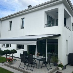 Growindo Dominik Tuszyński - Elegancka, rozłożona markiza tarasowa w kolorze szarym, przylegająca do białego domu z dużymi oknami. Na tarasie stół z krzesłami i dekoracje kwiatowe.