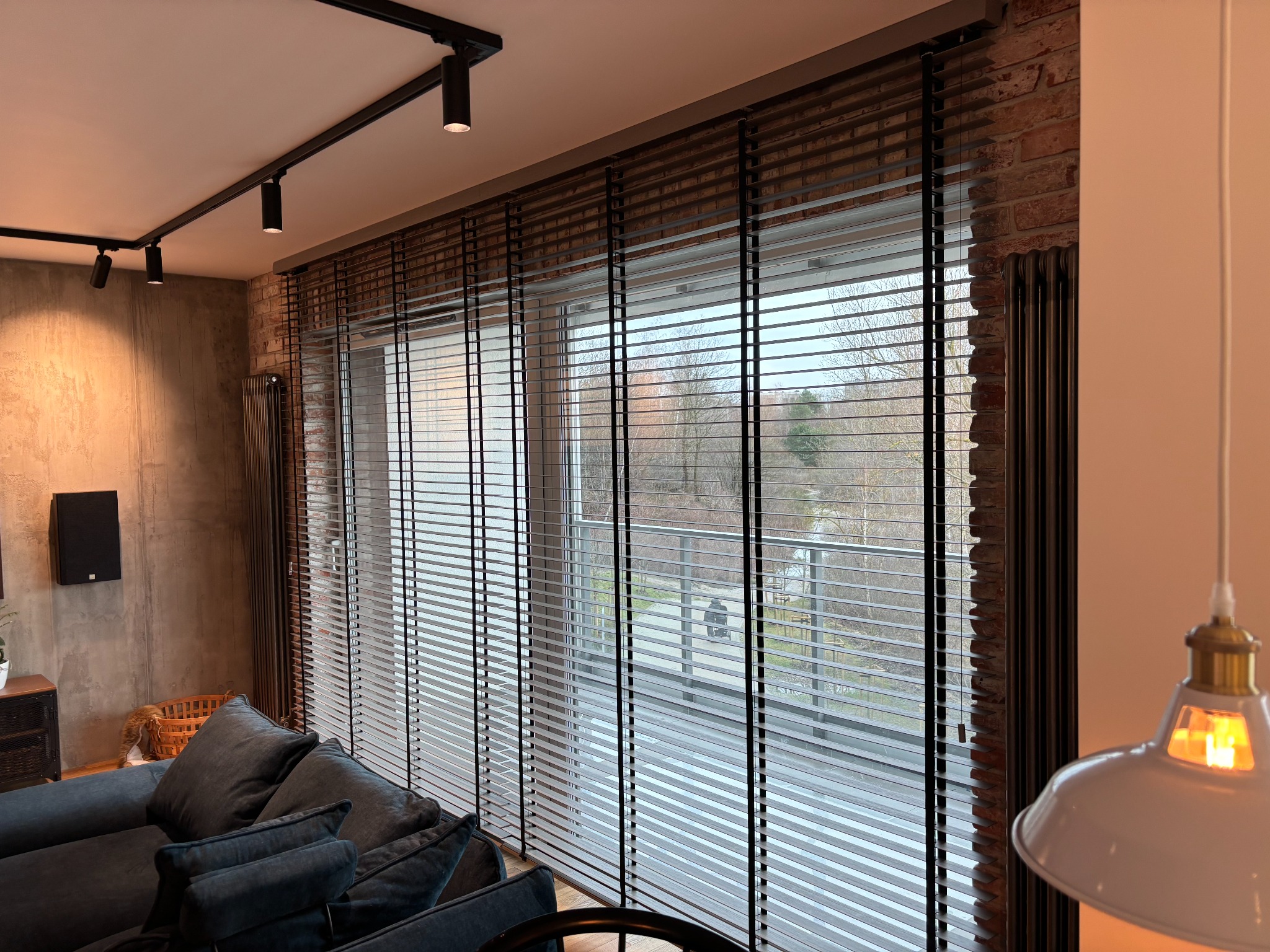 Wnętrze salonu z dużym oknem osłoniętym poziomymi żaluzjami drewnianymi w kolorze ciemnego brązu, widok na zewnątrz z drzewami i chodnikiem.