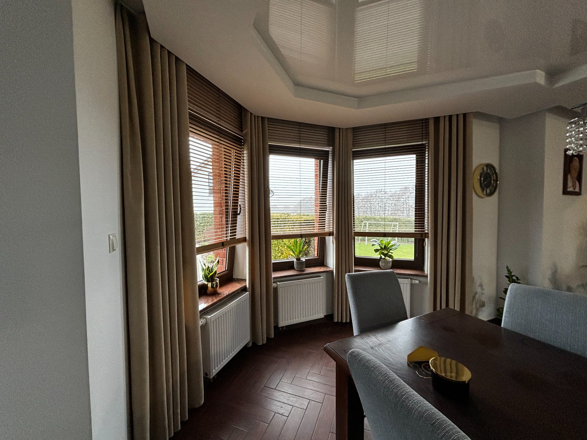 Wnętrze jadalni z trzema oknami osłoniętymi drewnianymi żaluzjami poziomymi i beżowymi zasłonami, na parapetach doniczki z roślinami, pod oknami białe grzejniki, ciemna podłoga ułożona we wzór...