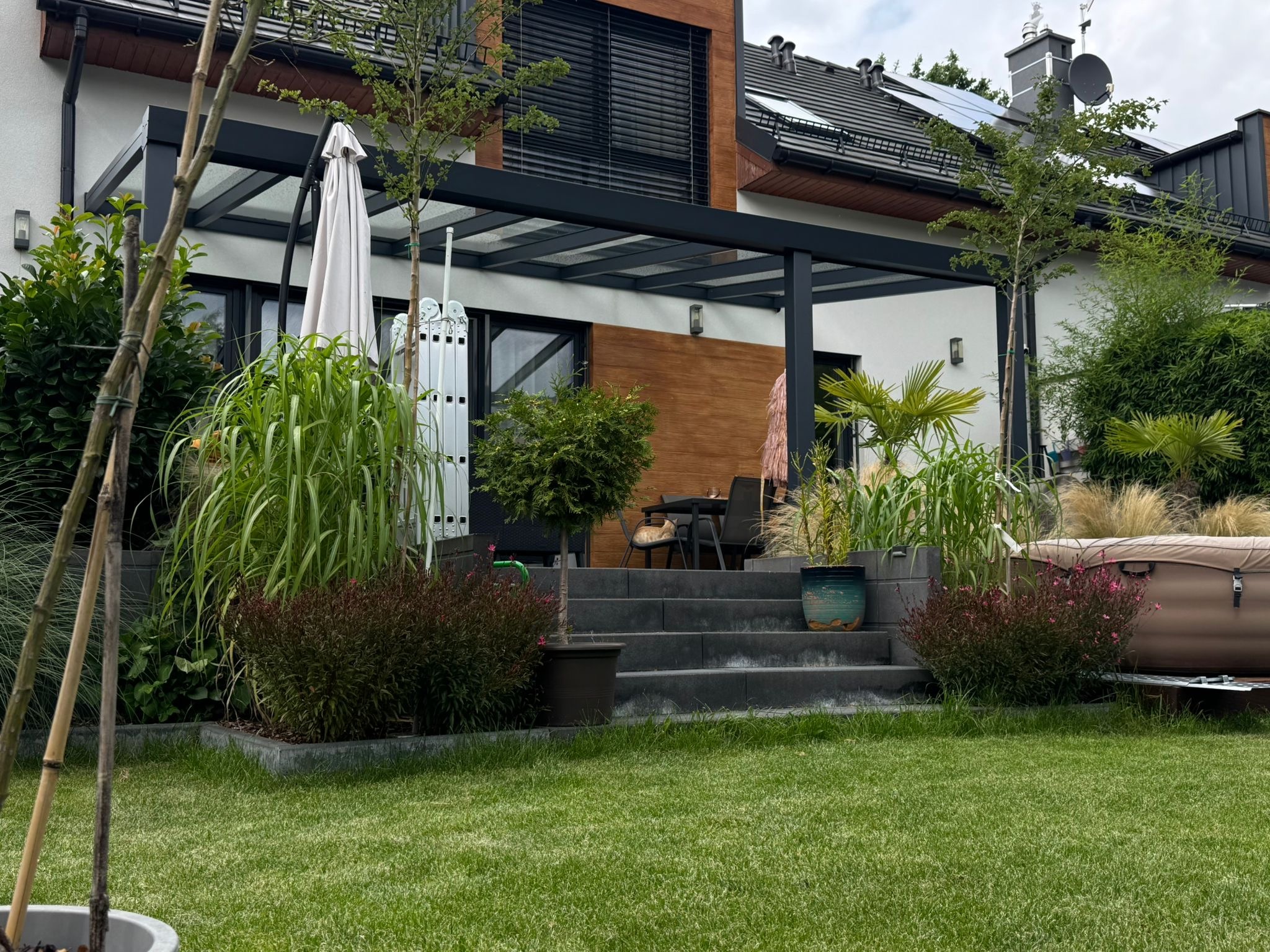 Nowoczesna pergola metalowa z zadaszeniem na tarasie domu jednorodzinnego, otoczona zielenią. Widoczne schody tarasowe i elementy wyposażenia ogrodu.
