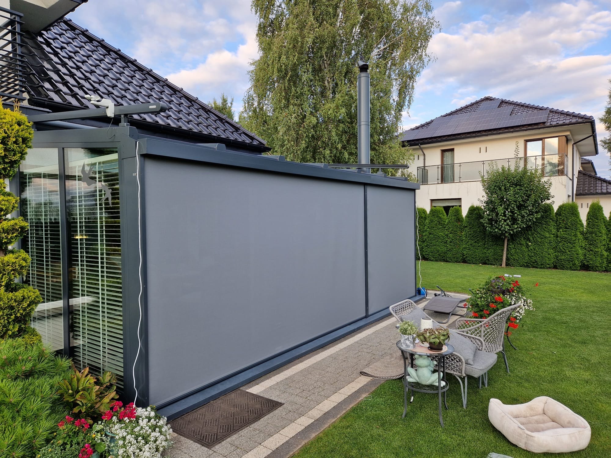 Szara, zwinięta osłona przeciwsłoneczna na tarasie z meblami ogrodowymi i zielonym trawnikiem. Widoczny fragment domu z panelami słonecznymi na dachu.