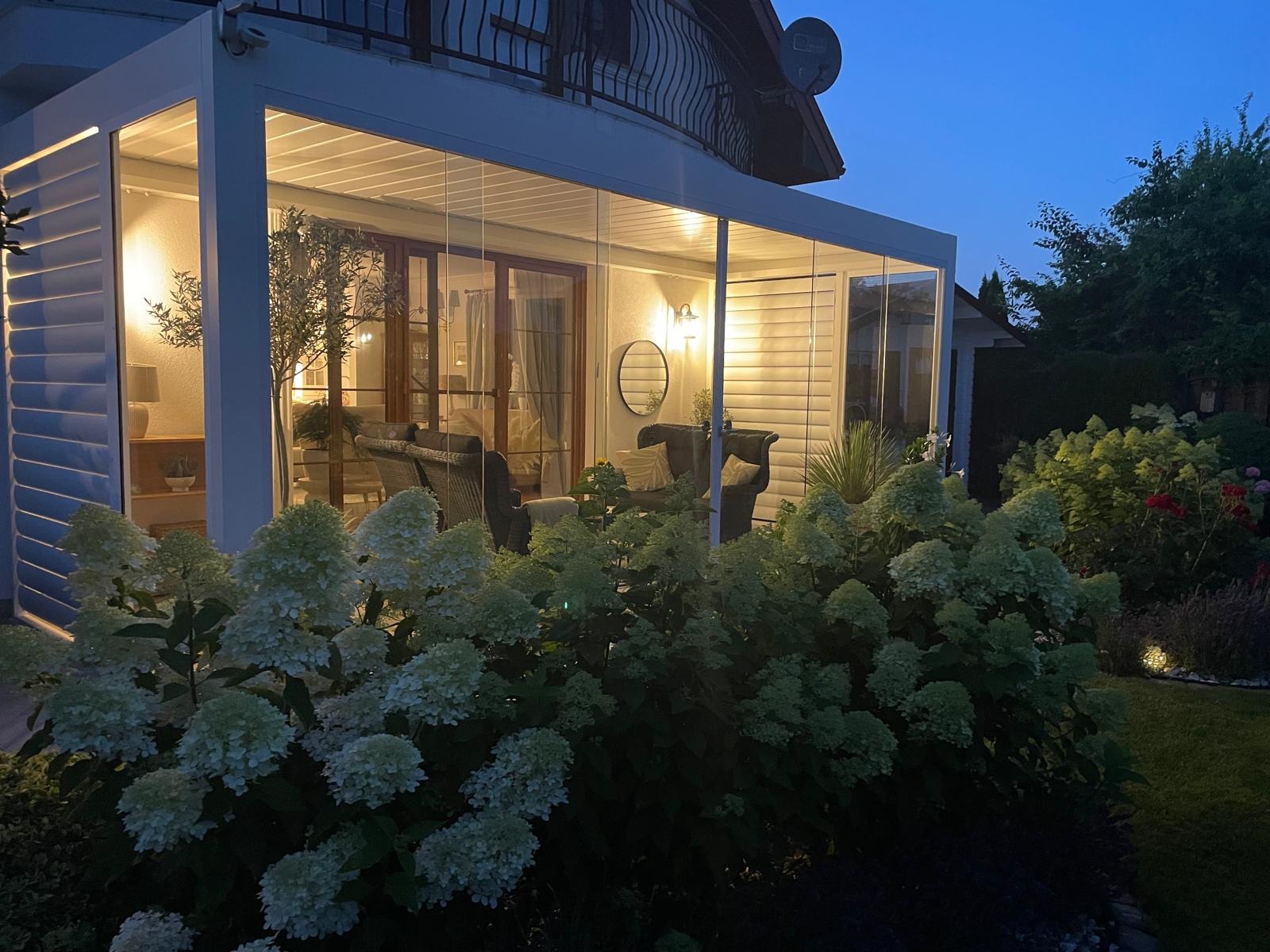 Szklana zabudowa tarasu z oświetleniem w Białogardzie, widok z ogrodu. Wnętrze z meblami wypoczynkowymi, na pierwszym planie kwitnące hortensje.