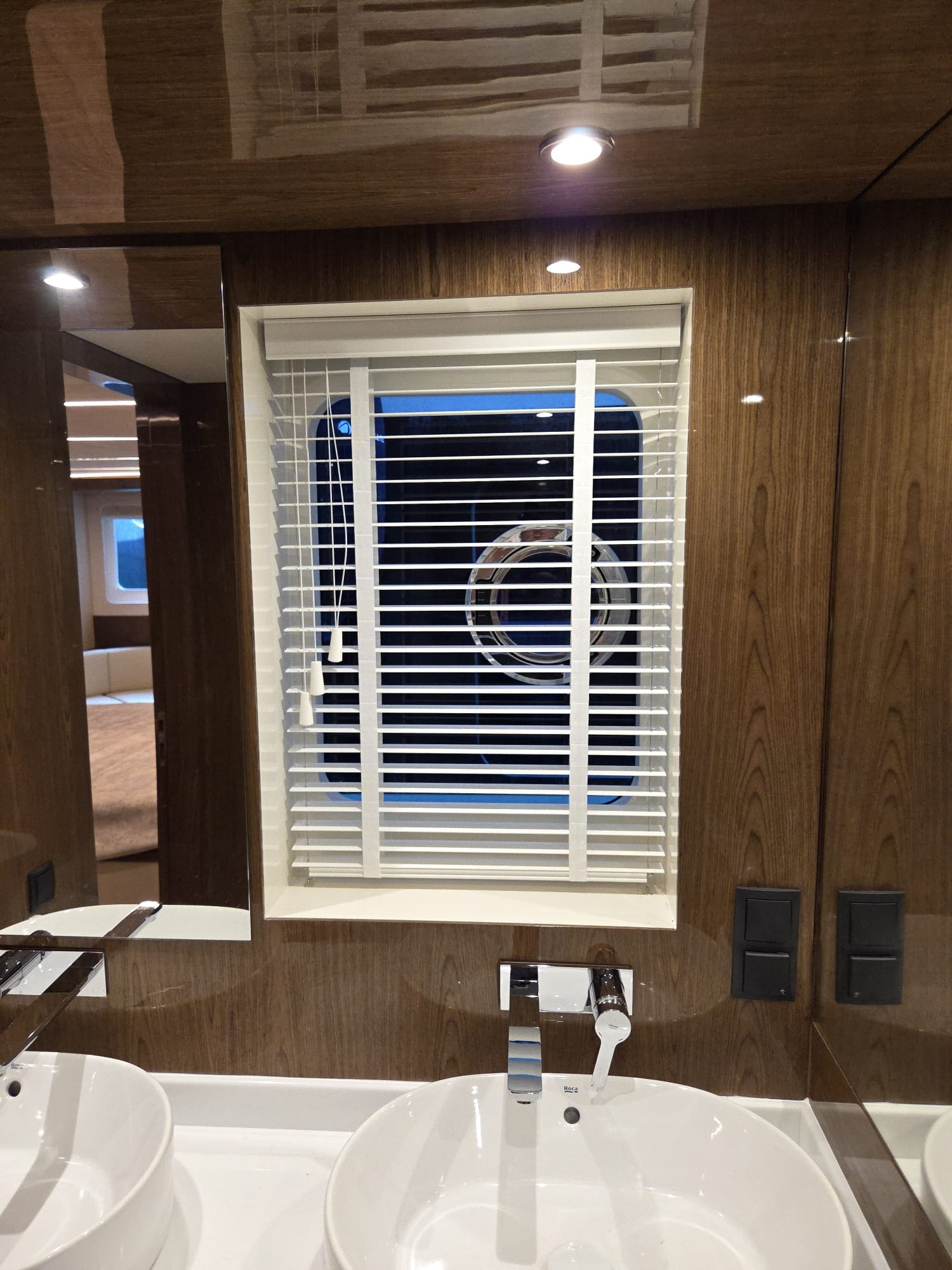 Białe żaluzje poziome w łazience jachtu, widoczne przez okno pralka. Umywalki i armatura na pierwszym planie, drewniane wykończenie ścian.