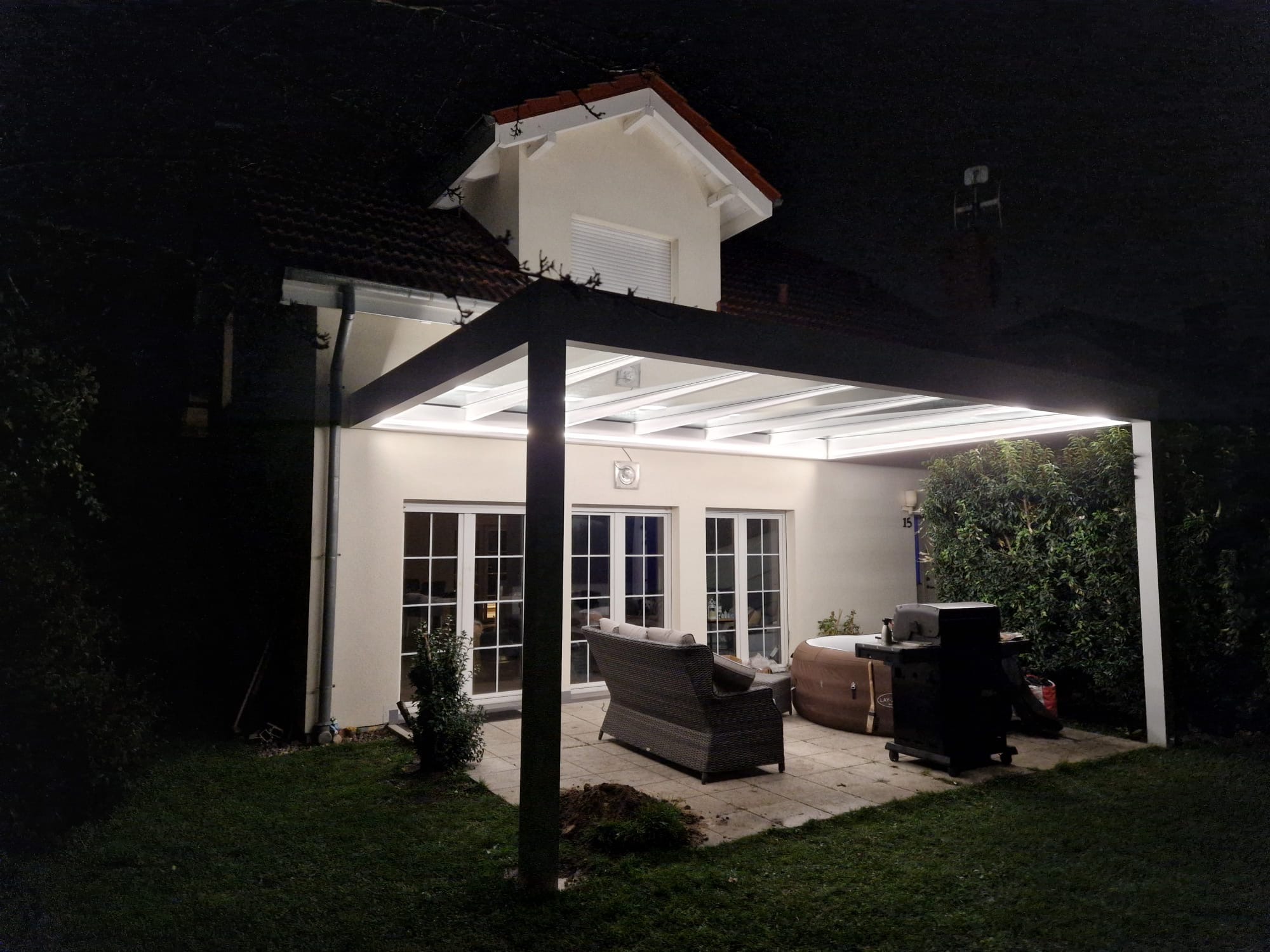 Oświetlona pergola z metalową konstrukcją i białym zadaszeniem, przylegająca do domu z oknami i roletą. Na tarasie meble ogrodowe i jacuzzi, nocne oświetlenie.