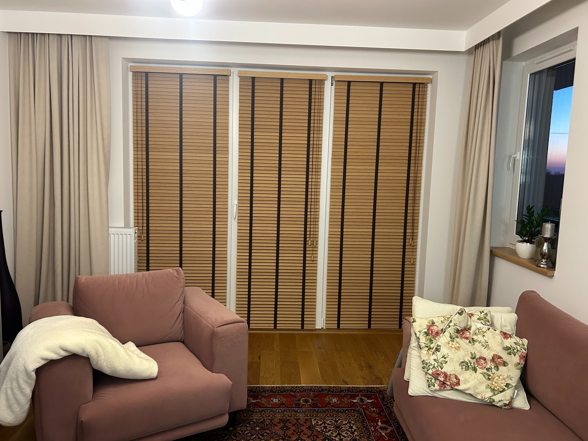 Salon z drewnianymi żaluzjami w oknach. Widoczne beżowe zasłony, różowy fotel i kanapa. Na parapecie rośliny doniczkowe. Ciepłe oświetlenie wnętrza.