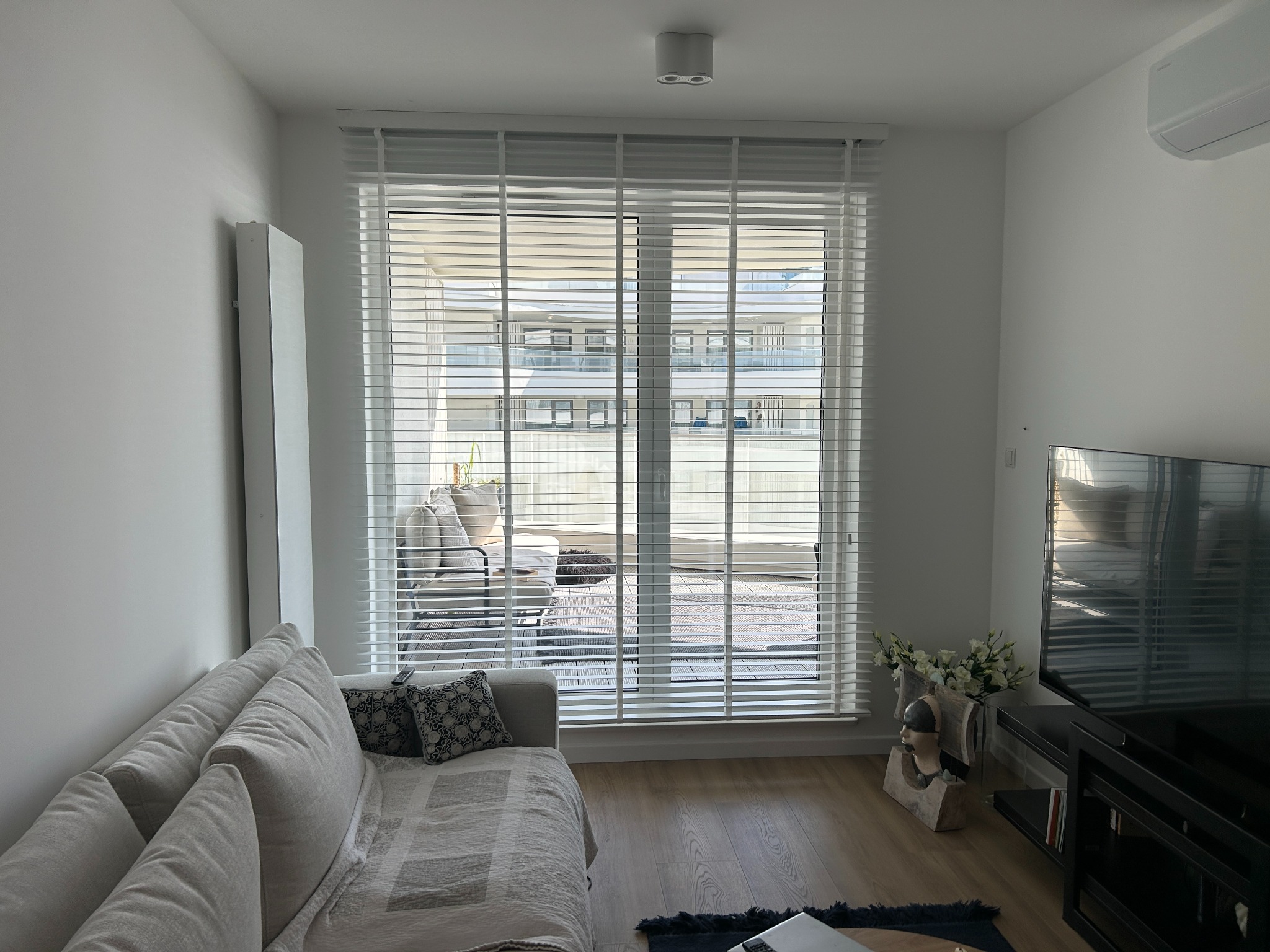 Jasne wnętrze z białymi żaluzjami poziomymi, przez które widać balkon z meblami wypoczynkowymi. Minimalistyczny design, naturalne światło.