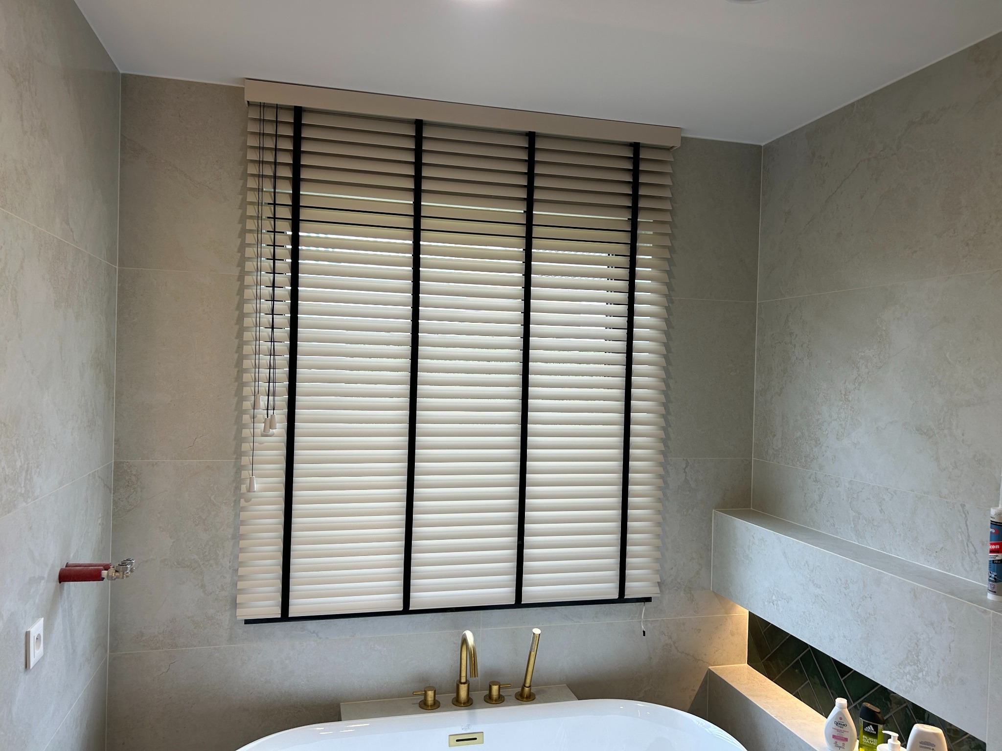 Beżowe poziome lamele z czarnymi taśmami w łazience z wanną i złotą armaturą. Stonowana kolorystyka płytek i oświetlenie tworzą spójny, elegancki wystrój.
