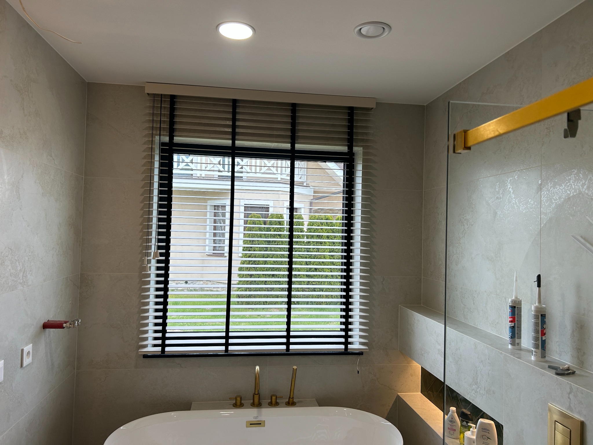 Beżowe poziome żaluzje z czarną ramą okienną w łazience z widokiem na dom i zieleń. Armatura łazienkowa w kolorze złotym. Widoczne tuby z silikonem.
