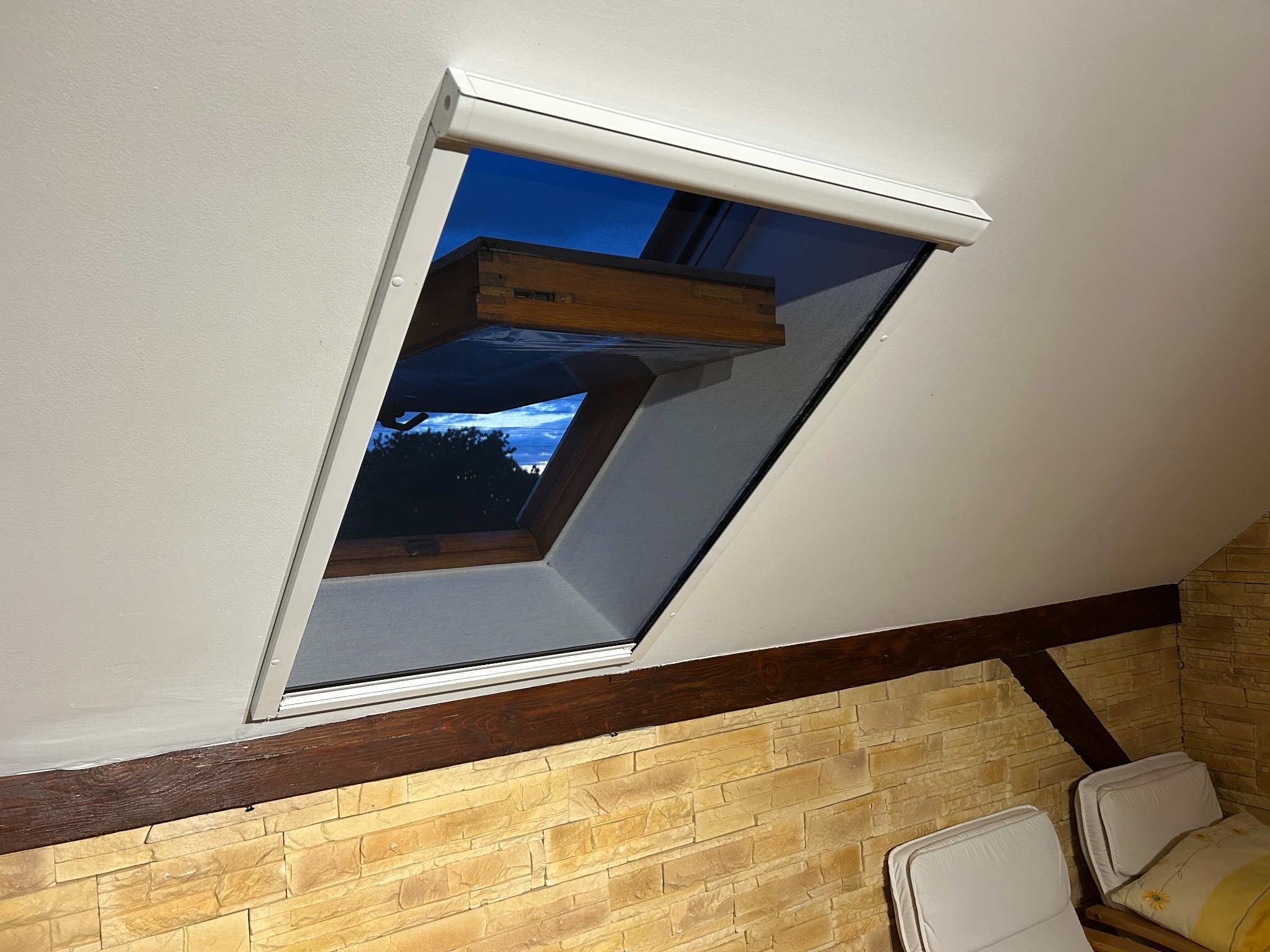 Okno dachowe z moskitierą w białej ramie, widok na wieczorne niebo. W tle ściana z kamienia dekoracyjnego i fragment mebli. Ujęcie od dołu, podkreślające montaż.