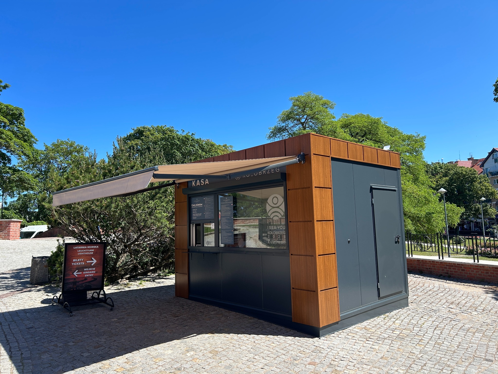 Kiosk biletowy z markizą w Kołobrzegu, z napisem 'KASA' i logo. Widoczny szyld informacyjny o Latarni Morskiej. Jasne niebo i bruk na pierwszym planie.