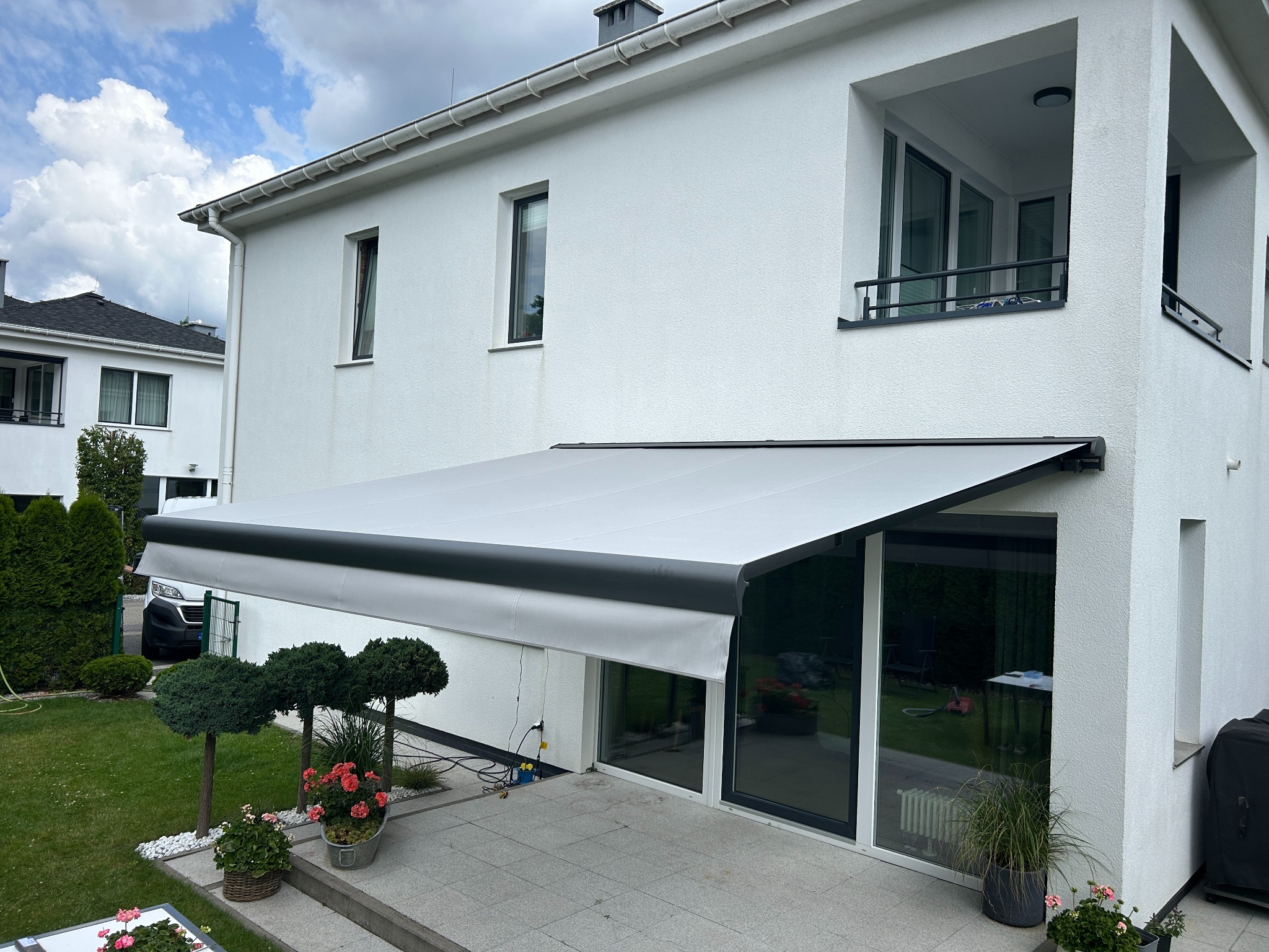 Biała markiza tarasowa z czarnym obramowaniem, zamontowana przy nowoczesnym domu z ogrodem. Widoczne rośliny ozdobne i fragment tarasu.