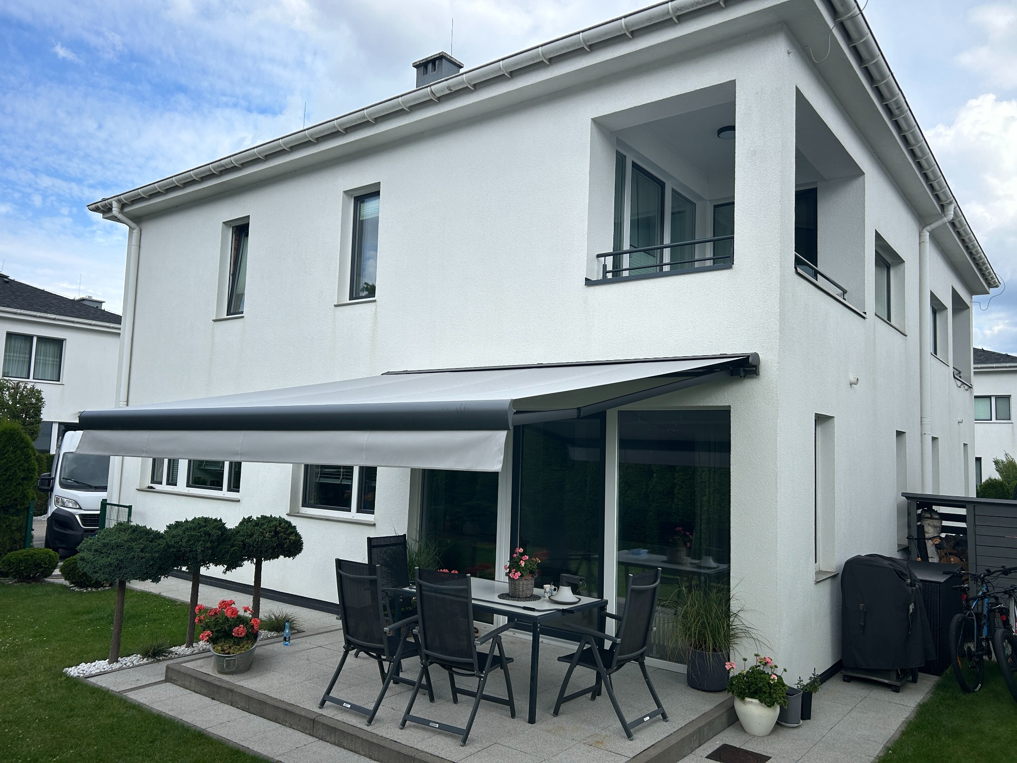 Elegancka, rozłożona markiza tarasowa w kolorze szarym, przylegająca do białego domu z dużymi oknami. Na tarasie stół z krzesłami i dekoracje kwiatowe.