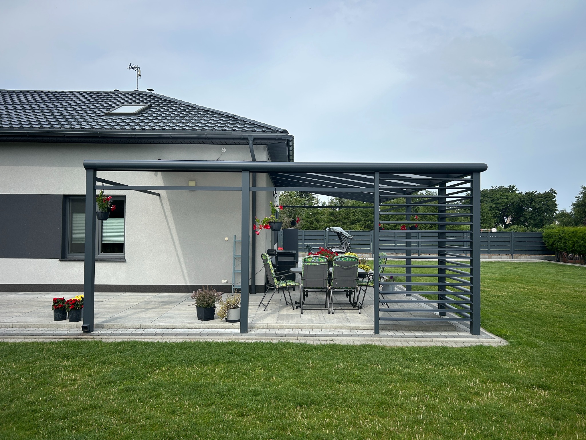 Nowoczesna, szara pergola metalowa przylegająca do domu, z widocznym zestawem mebli ogrodowych. Uwagę zwracają kwiaty w donicach i zadbany trawnik.