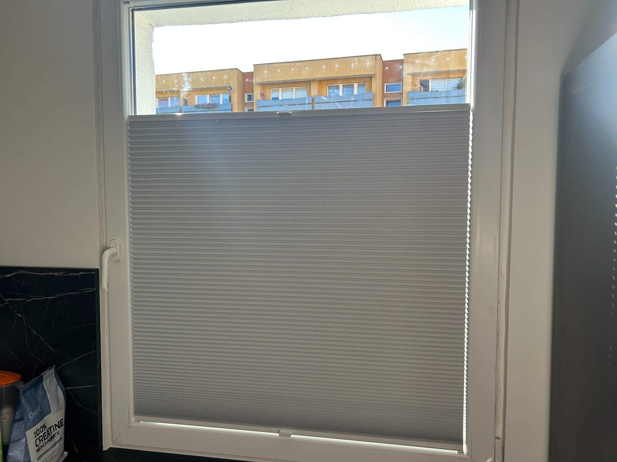 Szara plisa okienna zamontowana w białej ramie okiennej, z widokiem na budynki mieszkalne w tle. Widoczne elementy wyposażenia wnętrza po lewej stronie.