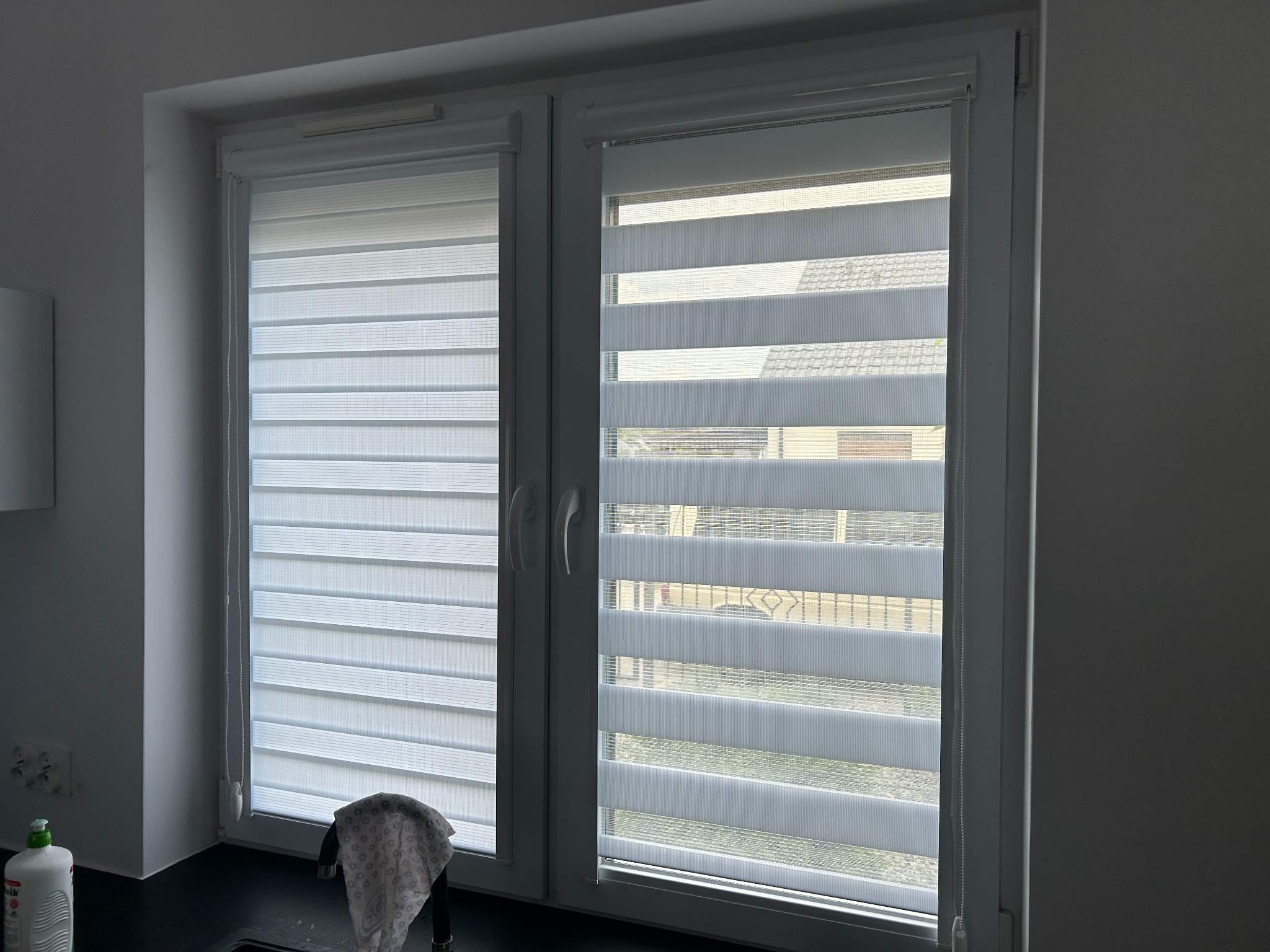 Okno z zamontowanymi, białymi roletami dzień-noc, widoczne z wnętrza pomieszczenia. Na parapecie leży szmatka. Widok na zewnątrz przez prześwity rolet.