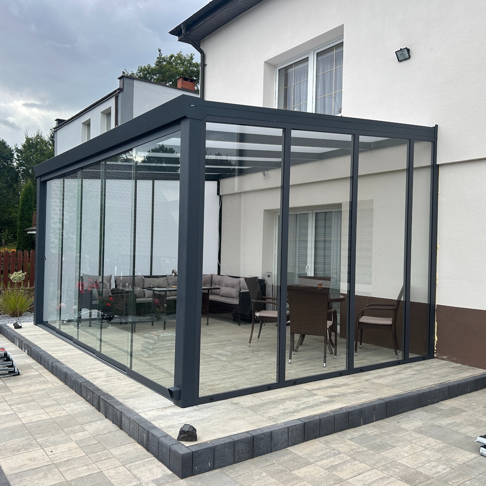 Szklana, nowoczesna pergola metalowa przylegająca do budynku. Wewnątrz widoczne meble ogrodowe. Szara konstrukcja, przeszklone ściany. Widok z zewnątrz, kostka brukowa.
