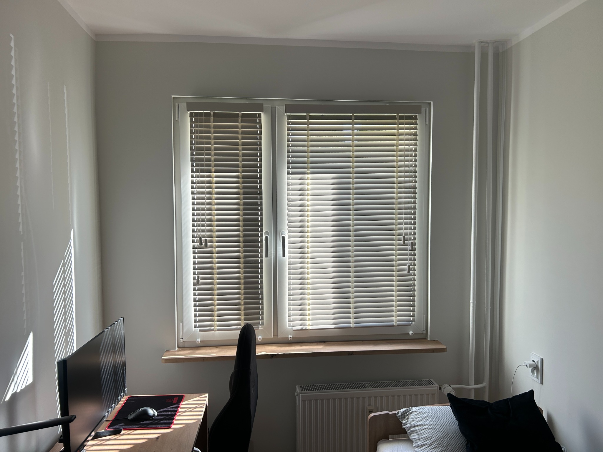 Jasne wnętrze pokoju z oknem, na którym zamontowano poziome, beżowe listwy. Na ścianie widoczne refleksy światła słonecznego.