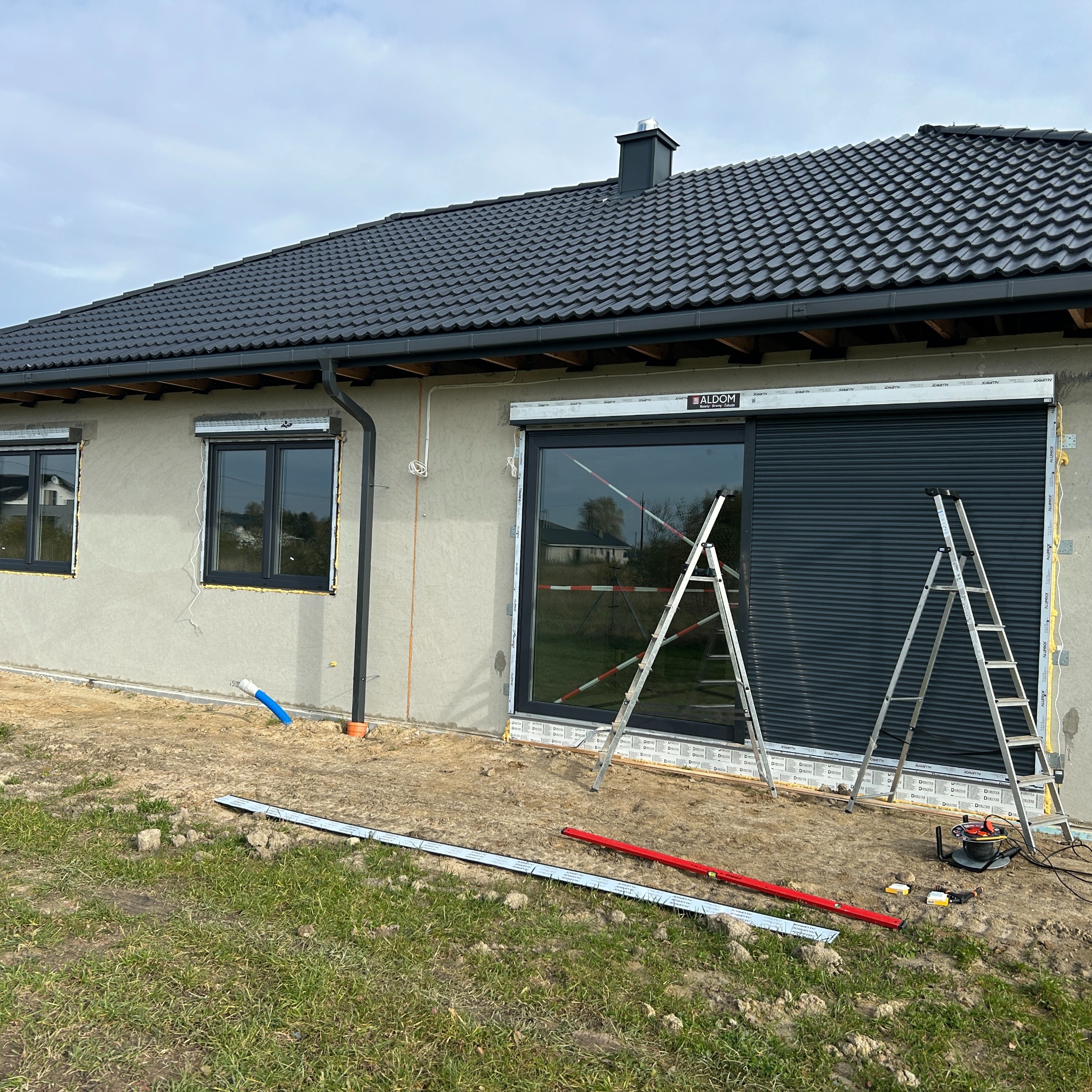 Montaż rolety zewnętrznej na oknie domu jednorodzinnego. Widoczne drabiny, narzędzia i częściowo opuszczona roleta w kolorze grafitowym. Prace montażowe w toku.