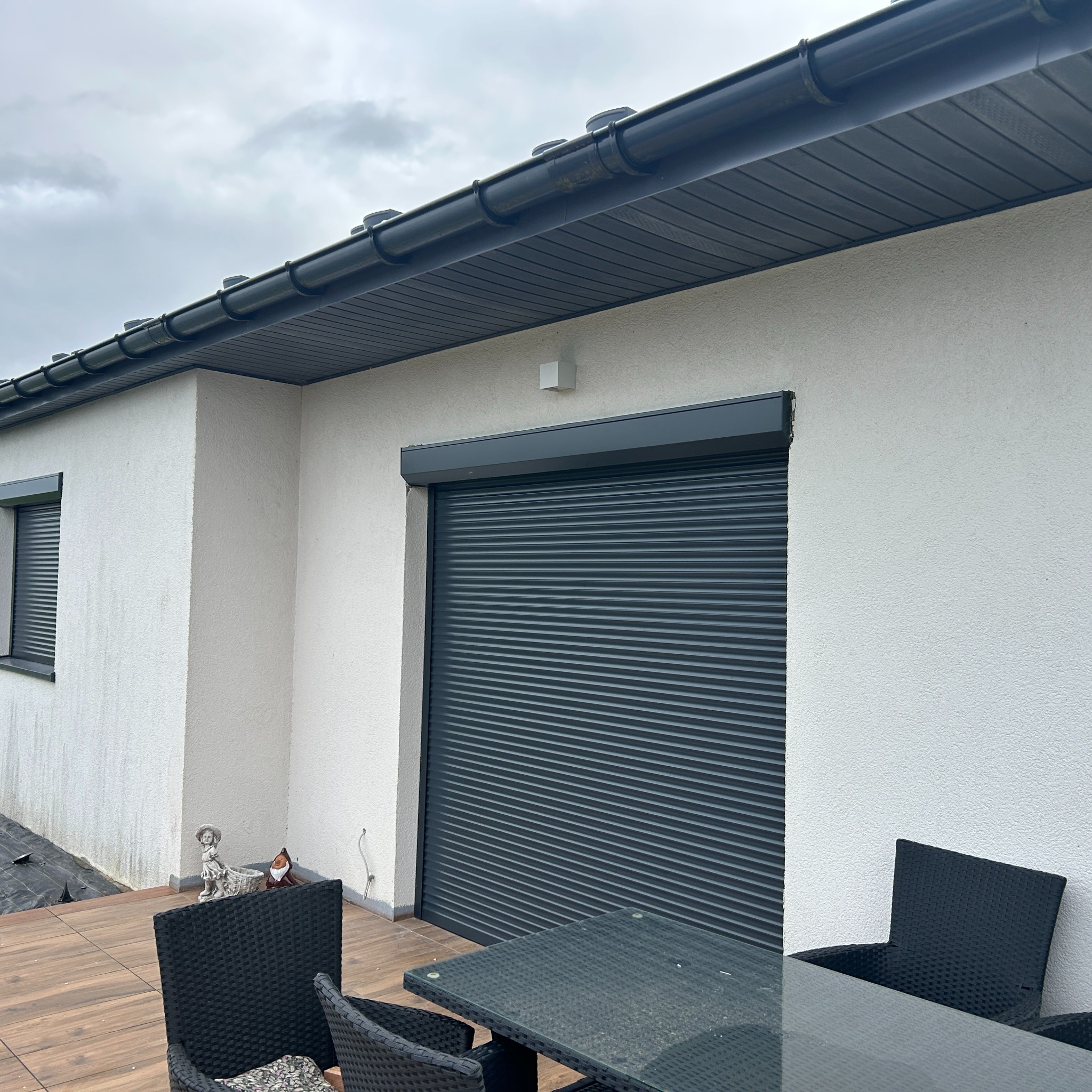 Elewacja domu z zamkniętą, szarą roletą zewnętrzną na oknie tarasowym. Widoczne meble ogrodowe i dekoracje. Pochmurne niebo w tle.