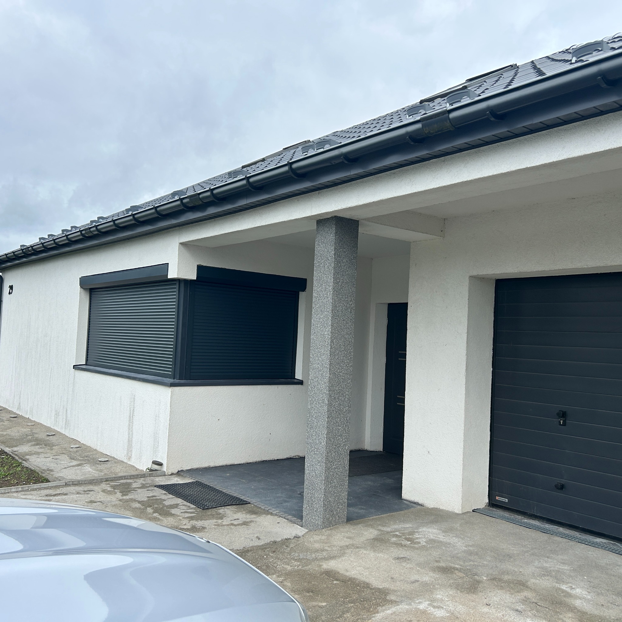 Nowoczesny dom z szarą stolarką okienną i garażową. Elewacja biała, dachówka grafitowa. Widoczny fragment srebrnego samochodu. Pochmurne niebo.