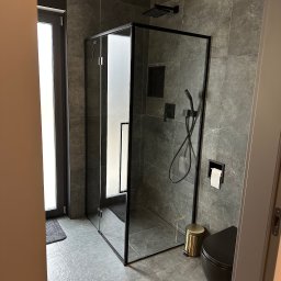 BUDEXPERTS IHOR VOLOVYK - Nowoczesna łazienka z szarymi płytkami imitującymi kamień, kabiną prysznicową z czarnymi ramami, czarną toaletą wiszącą i złotym koszem na śmieci.