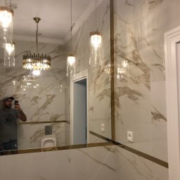 BUDEXPERTS IHOR VOLOVYK - Elegancka łazienka wykończona płytkami imitującymi marmur z złotymi akcentami, widoczne wiszące lampy i odbicie osoby robiącej zdjęcie.