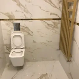 Nowoczesna toaleta z otwartą klapą, wykończona płytkami imitującymi marmur z złotymi akcentami i ozdobnym grzejnikiem drabinkowym w kolorze złota.