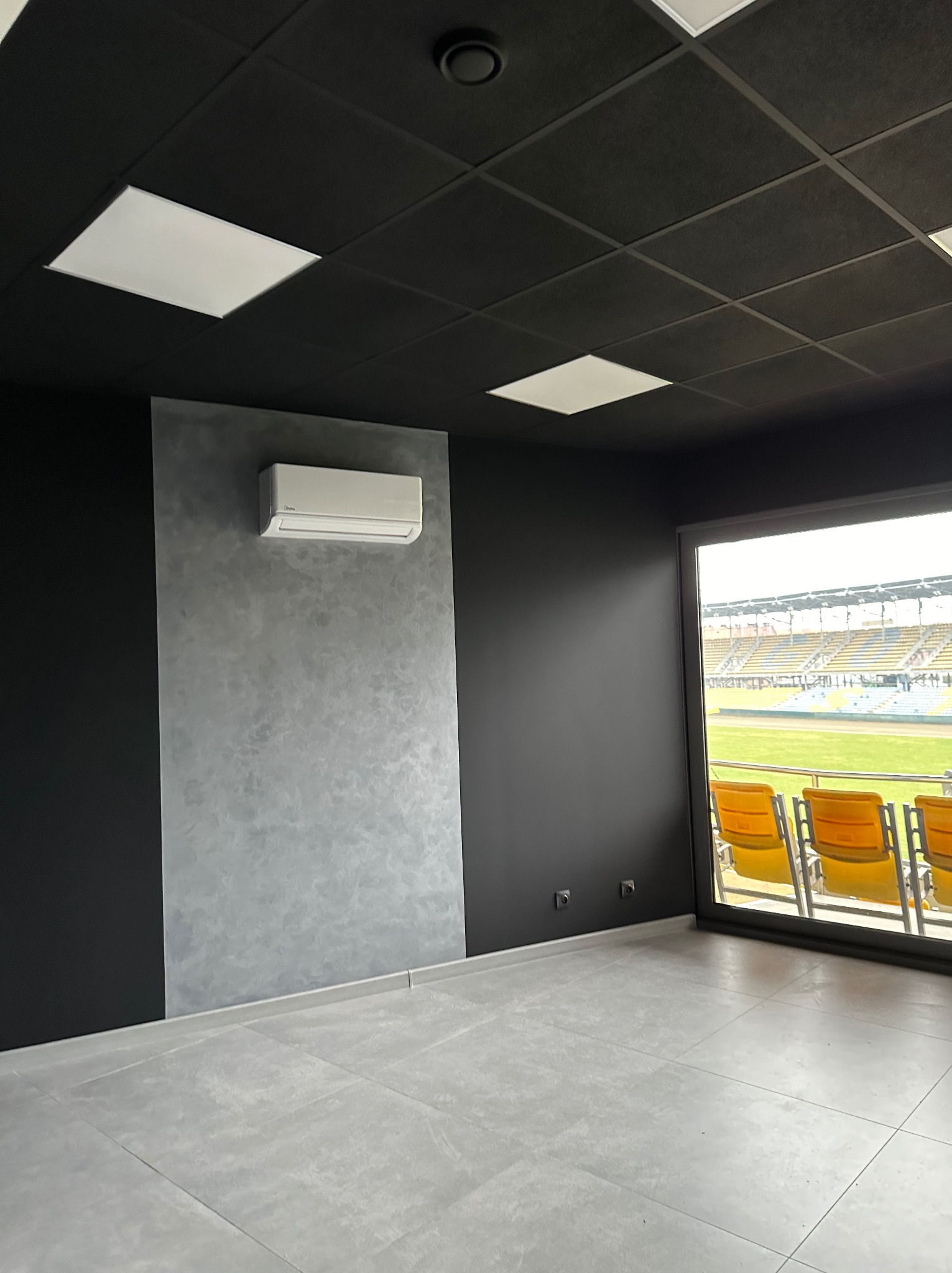 Wnętrze pomieszczenia z widokiem na stadion: czarne ściany i sufit z kwadratowymi lampami, ściana akcentowa z szarą fakturą, biały klimatyzator, szare płytki podłogowe, okno z widokiem na trybuny...
