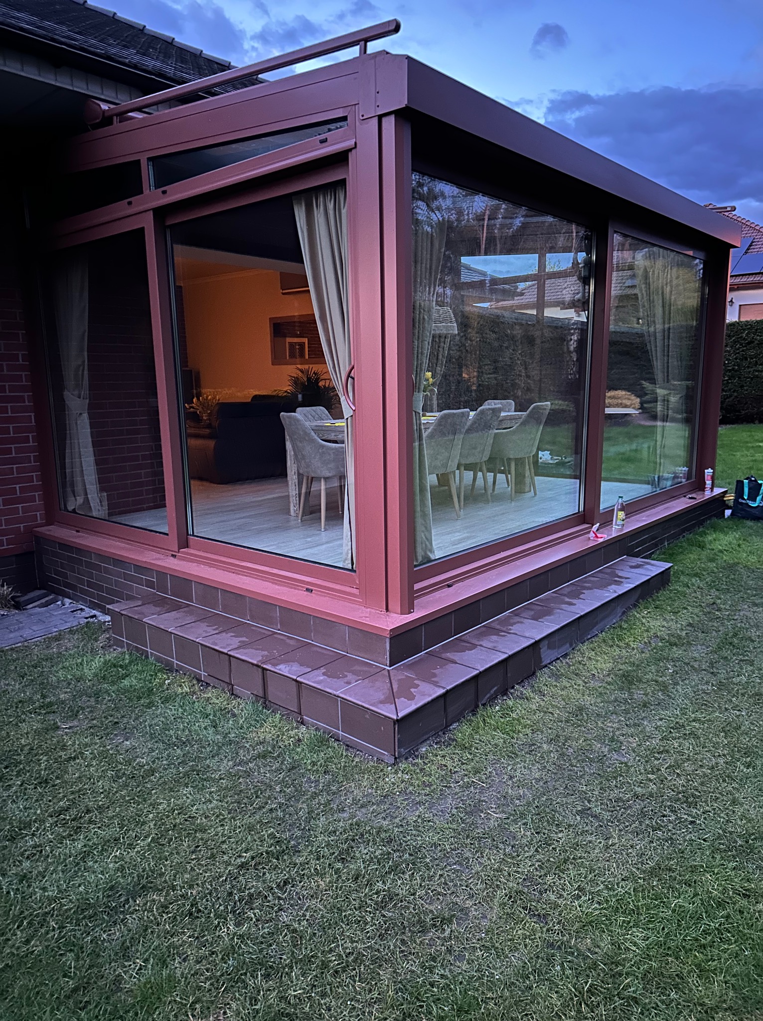 Nowoczesna, przeszklona weranda z brązowymi ramami, widok z zewnątrz na stół i krzesła wewnątrz, brązowe płytki na schodach wejściowych, zielony trawnik.